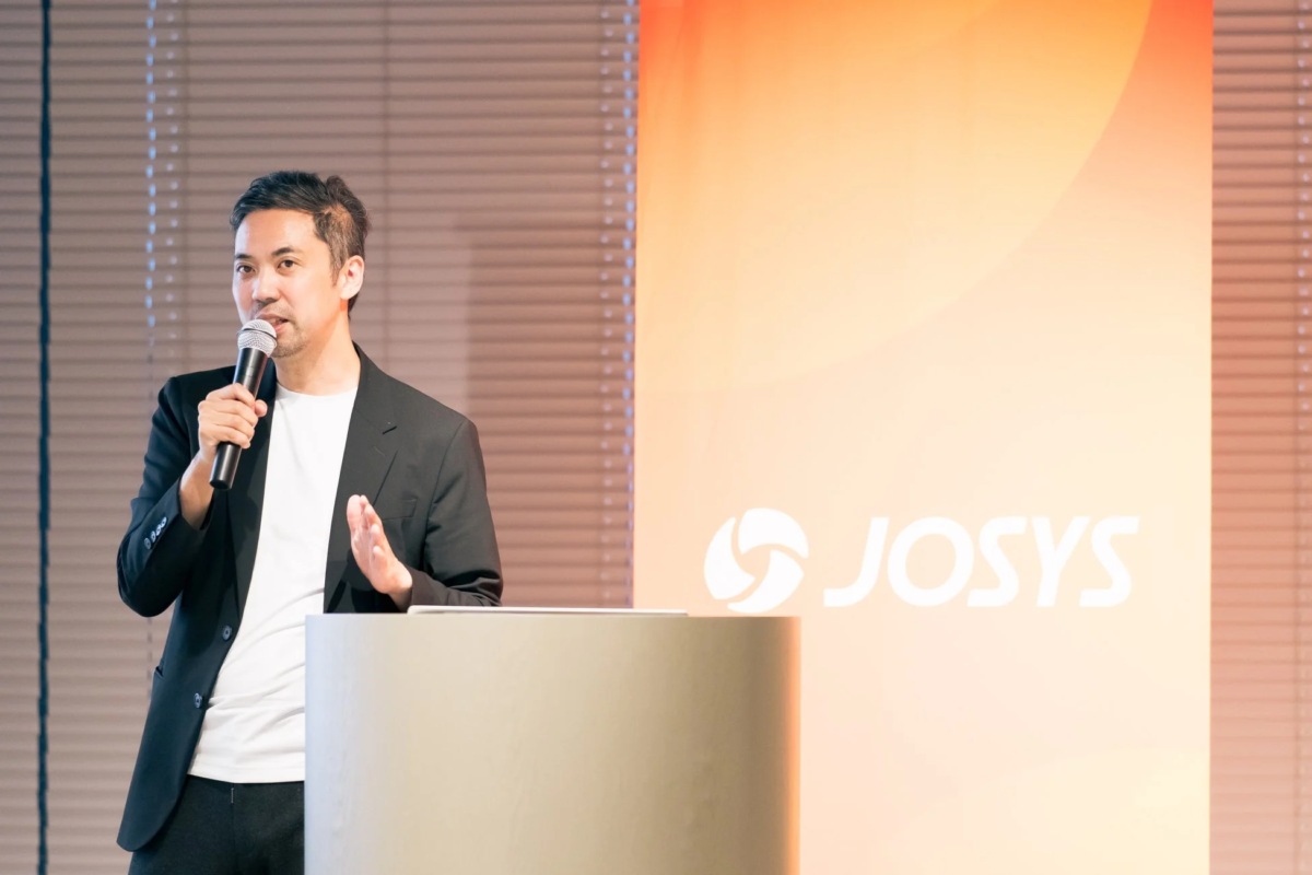 ビジネスイベントで講演を行う男性の様子。マイクを手に、壇上で話しており、背景には「JOSYS」のロゴが見えます。