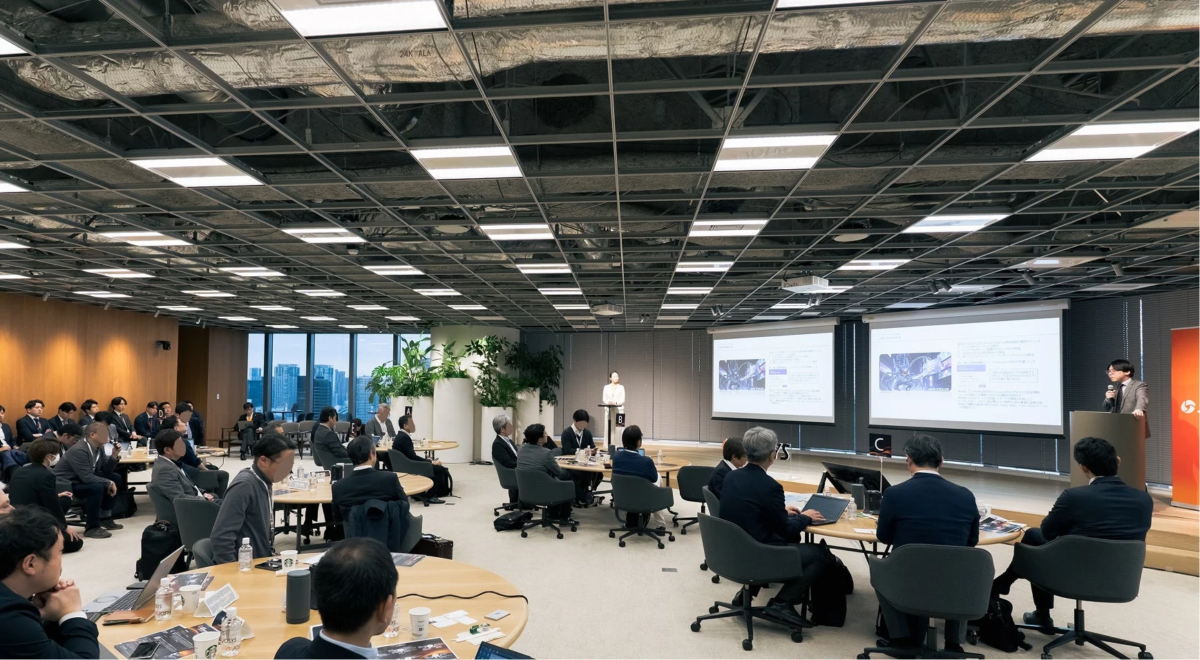 現代的なオフィス空間でビジネスプレゼンテーションが行われています。講演者が壇上で話し、多数の参加者が丸テーブルに着席して熱心に耳を傾けています。