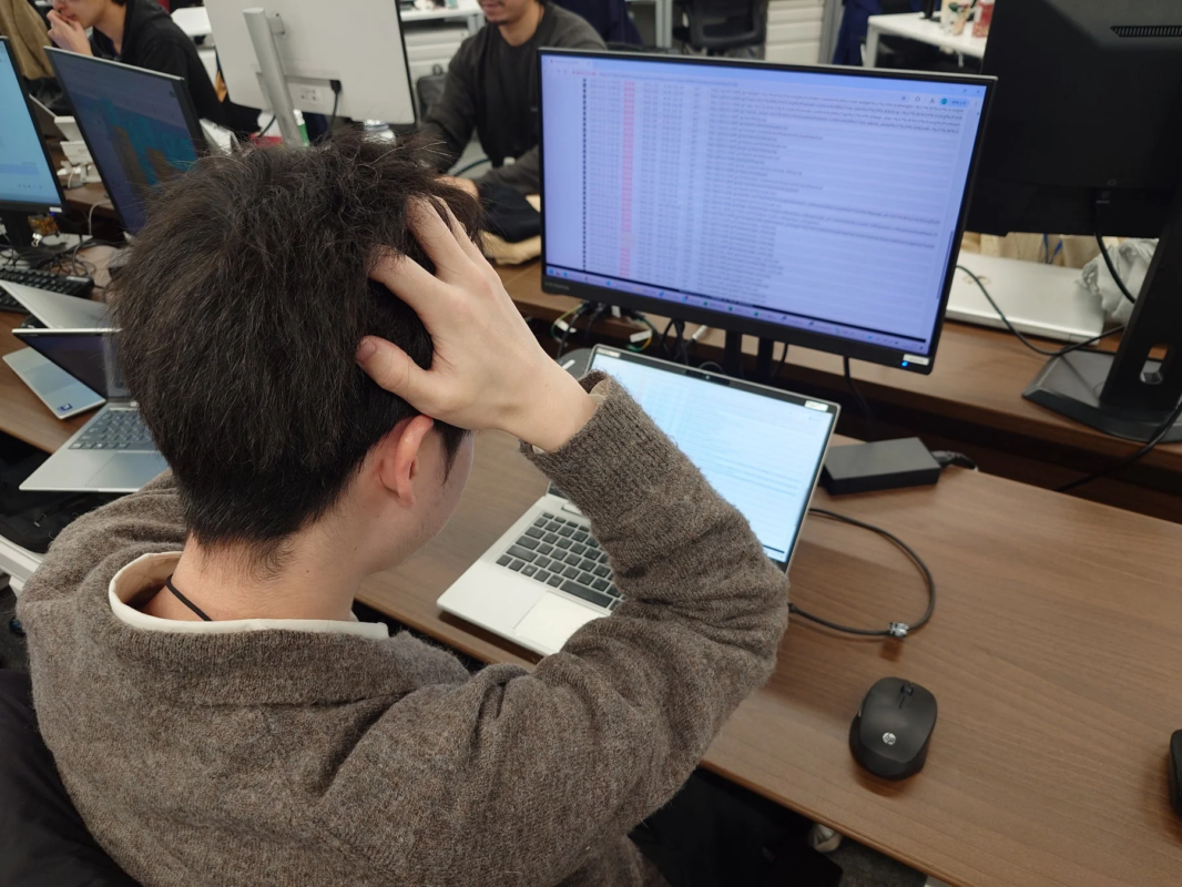 オフィスでパソコンに向かう人物の後ろ姿。複数のモニターを見つめ、片手を頭に当てて何かに悩んでいるか、深く集中している様子