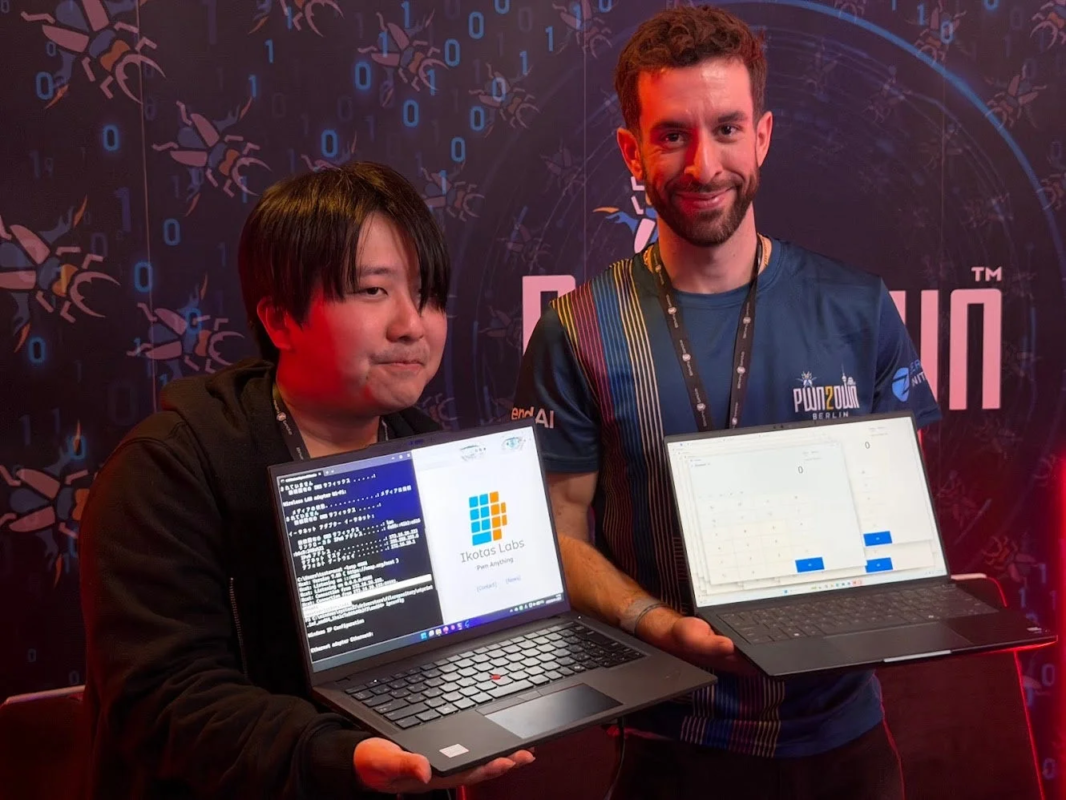 Pwn2Own Berlinのイベントで、二人の男性がラップトップを掲げて笑顔を見せている
