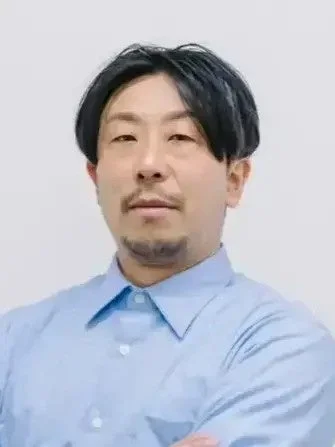 水色のシャツを着た東洋系の男性がカメラをまっすぐ見つめているポートレート写真です。ひげと口ひげがあり、黒い短髪が特徴的です。