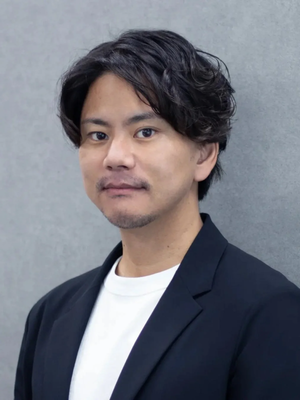 濃い色のジャケットと白いTシャツを着用し、無精髭を生やしたアジア人男性のバストアップポートレート。グレーの背景を背に、カメラをまっすぐ見つめている。