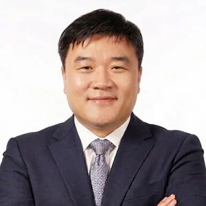 紺色のスーツとネクタイを着用したアジア系の男性が、白い背景の前で腕を組み、カメラに向かって微笑んでいる上半身のポートレート写真です。