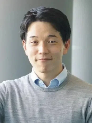 水色のシャツとグレーのセーターを着用した短髪のアジア人男性が、カメラに向かって穏やかに微笑んでいる上半身のポートレートです。