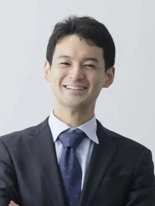 白い背景の前で、スーツを着た男性が笑顔でカメラを見ているポートレート写真です。ビジネスシーンや公式なプロフィール写真に適しています。
